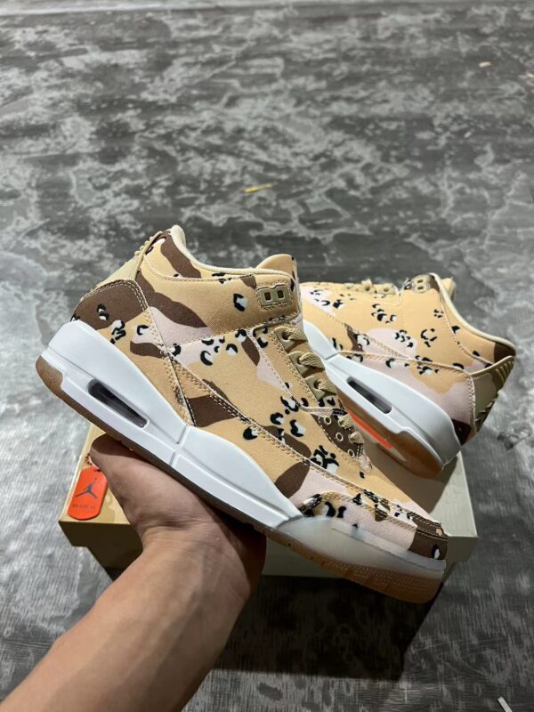 A pair of Jordan 'Desert Camo' lifestyle sneakers, displaying their camouflage pattern, white midsoles with visible Air units, and gum outsoles, resting on a beige shoe box.