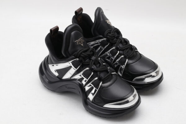 A single black chunky athletic sneaker with a large, sculptural wavy black sole, featuring reflective silver metallic overlays on the upper and black laces, displayed from a side profile against a plain white background.