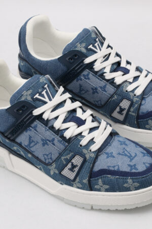 A pair of blue denim-textured low-top sneakers with a subtle all-over monogram pattern, prominent white laces, a white outsole, and visible blue side details, displayed from a top-down angle on a light grey background.