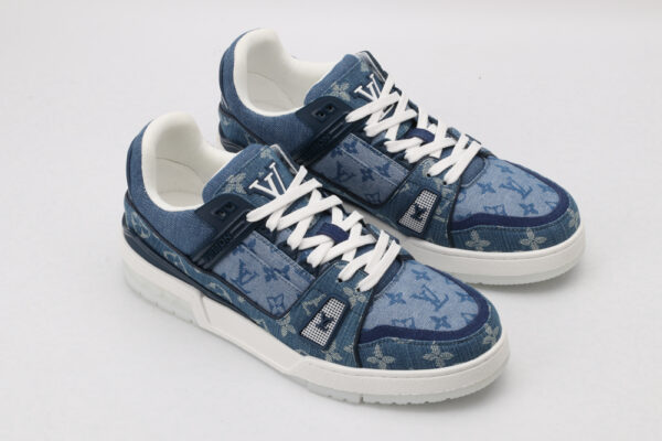 A pair of blue denim-textured low-top sneakers with a subtle all-over monogram pattern, prominent white laces, a white outsole, and visible blue side details, displayed from a top-down angle on a light grey background.