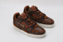 A pair of brown and tan monogrammed low-top sneakers with darker brown accents, brown laces, and a contrasting white outsole, displayed against a light grey background.