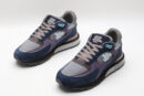 A pair of athletic sneakers in navy blue suede, purple fabric, and grey mesh, featuring taupe laces, light blue accents, and a layered brown and grey sole with branding on the side, displayed against a plain white background
