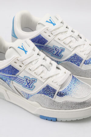 A pair of white low-top sneakers heavily embellished with shimmering silver rhinestones across the toe and sides, featuring bold blue rhinestone accents forming a logo and geometric patterns, set against a plain white background.