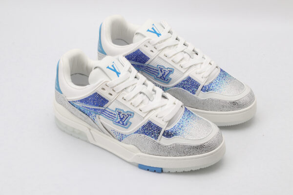 A pair of white low-top sneakers heavily embellished with shimmering silver rhinestones across the toe and sides, featuring bold blue rhinestone accents forming a logo and geometric patterns, set against a plain white background.