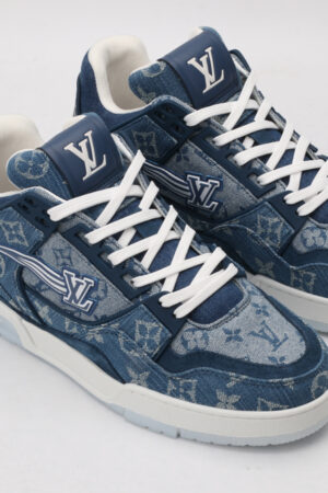 A pair of blue denim-textured low-top sneakers with a subtle all-over monogram pattern, prominent white laces, a white outsole, and a blue side logo, displayed against a light grey background.