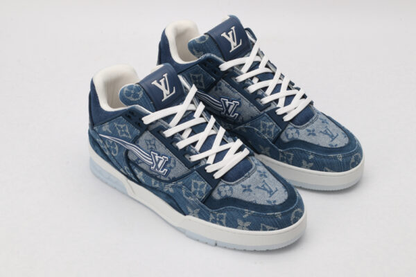 A pair of blue denim-textured low-top sneakers with a subtle all-over monogram pattern, prominent white laces, a white outsole, and a blue side logo, displayed against a light grey background.