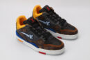 A pair of multi-toned low-top sneakers featuring brown and black monogrammed sections, vibrant blue accents on the sides, bright yellow interior lining, black laces, and a white outsole with subtle red detailing, displayed against a light grey background.