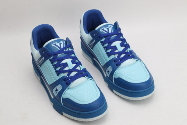 A pair of two-tone blue low-top sneakers, featuring darker blue overlays and lighter blue panels on the toe box and side, with matching blue laces, a white outsole, and intricate blue sole patterns, displayed against a light grey background.