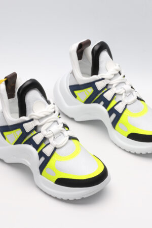 A pair of white chunky athletic sneakers with bold navy blue and neon yellow overlays, white laces, and a large, sculpted white sole with black accents, displayed against a light grey background.