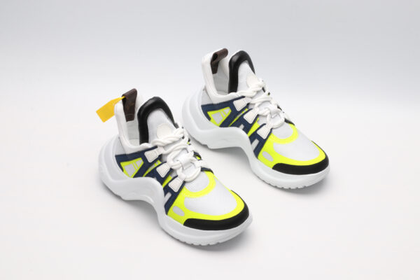 A pair of white chunky athletic sneakers with bold navy blue and neon yellow overlays, white laces, and a large, sculpted white sole with black accents, displayed against a light grey background.