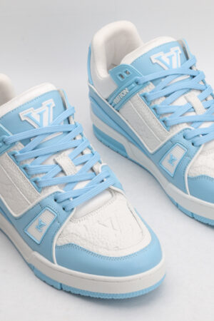 A pair of Louis Vuitton LV Trainer Low-Top Sneakers in white and light blue, viewed from an elevated angle, with the left shoe on its side showing the outsole with "VUITTON" branding.