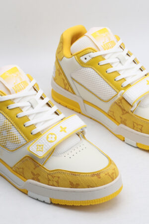 A pair of Louis Vuitton LV Trainer sneakers in yellow and white, featuring a white strap with monogram details, mesh panels, and white laces, shown from a top-down front view.