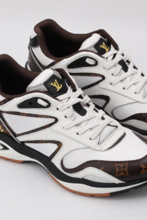 A pair of white athletic sneakers with brown monogram patterns on the toe and side, black outlining, white laces, and a multi-toned outsole with visible "VUITTON" branding on the heel, displayed against a light grey background