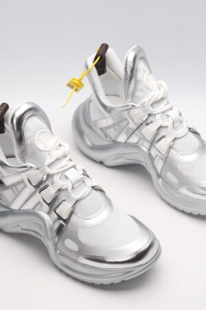 A pair of futuristic athletic sneakers featuring white mesh, sculpted metallic silver overlays, white laces, and a large, wavy metallic silver sole, displayed against a light grey background.