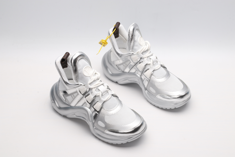 A pair of futuristic athletic sneakers featuring white mesh, sculpted metallic silver overlays, white laces, and a large, wavy metallic silver sole, displayed against a light grey background.