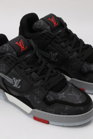 A pair of black and grey monogrammed low-top sneakers with red logo detailing on the tongue and a red accent on the outsole, featuring a white side logo and black laces, displayed against a light grey background.