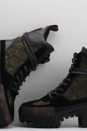 pair of dark green quilted high-top boots with black leather and suede panels, brown monogram details on the toe and base, black laces, and a chunky black lug sole, displayed against a light grey background.