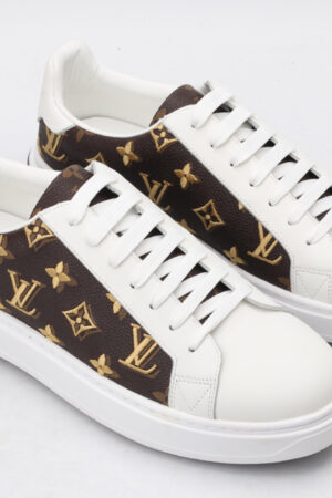 A pair of low-top sneakers with a brown and gold monogram pattern on the sides and white leather on the toe, tongue, and heel. The shoes have white laces and a thick white rubber sole, and are shown from a slightly elevated side angle on a clean white background.