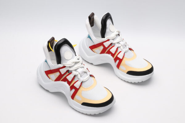 A pair of white chunky athletic sneakers with vibrant orange and light grey overlays, white laces, black accents on the toe and heel, and a large, sculpted white sole, displayed against a light grey background with one shoe showing the sole.