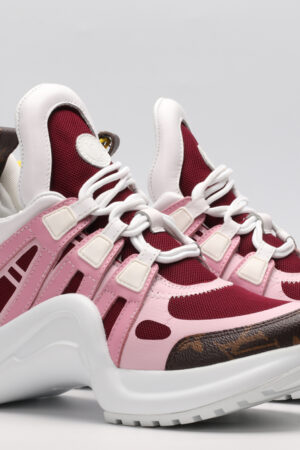 A pair of white chunky athletic sneakers with light pink and burgundy overlays, white laces, brown monogram accents on the toe, and a large, sculpted white sole, displayed against a light grey background.