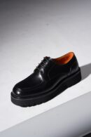 A pair of black polished leather derby shoes with a pronounced, chunky black rubber lug sole and vibrant orange interior lining, photographed on a white background with a shadow.
