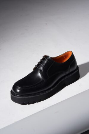 A pair of black polished leather derby shoes with a pronounced, chunky black rubber lug sole and vibrant orange interior lining, photographed on a white background with a shadow.