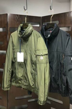 Moncler-style olive green lightweight windbreaker/utility jacket with a high collar, multiple zippered and flap pockets, and a circular logo patch on the left arm pocket.