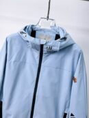 Close-up of a Moncler Grimm-style windbreaker jacket in black and light blue showing the drawstring hood, zipper, and signature logo patch.