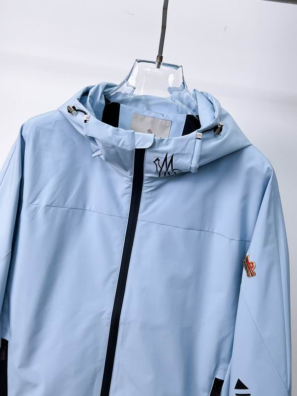 Close-up of a Moncler Grimm-style windbreaker jacket in black and light blue showing the drawstring hood, zipper, and signature logo patch.