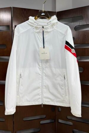 White Moncler Grimpeurs windbreaker jacket with a hood, front zipper, and red and black striped band on the left sleeve hanging on a gold hanger.