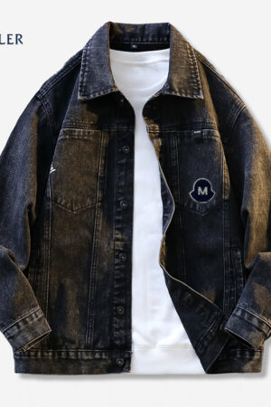 A dark wash black or indigo denim jacket in a trucker style, featuring a collar, button front, and a large, round black and white 'M' logo patch on the left chest pocket.