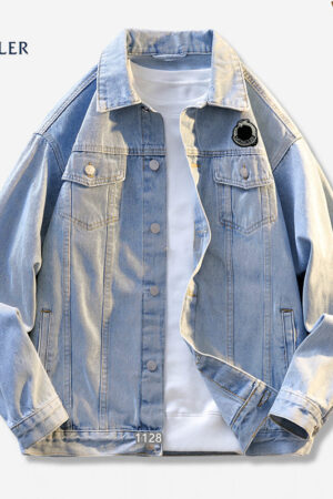 A light wash blue denim jacket in a trucker style, featuring a collar, button front, and a small, round black and white logo patch on the left chest pocket.
