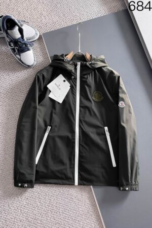 Close-up of an olive green hooded jacket featuring a white center zipper, an embroidered chest patch, and a signature logo on the sleeve.