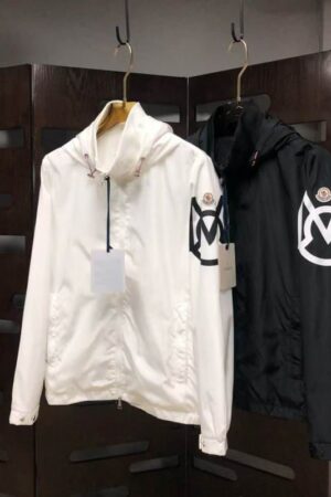Two hooded Moncler style windbreaker jackets hanging on display, one in white and one in black, both featuring a large, distinctive logo graphic patch on the left sleeve.