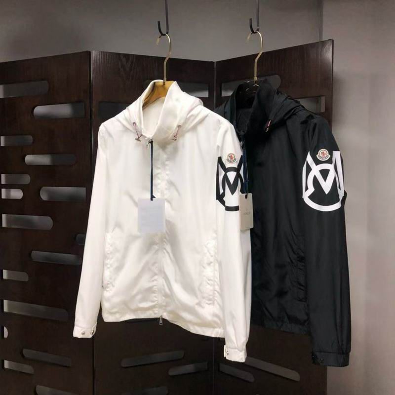Two hooded Moncler style windbreaker jackets hanging on display, one in white and one in black, both featuring a large, distinctive logo graphic patch on the left sleeve.
