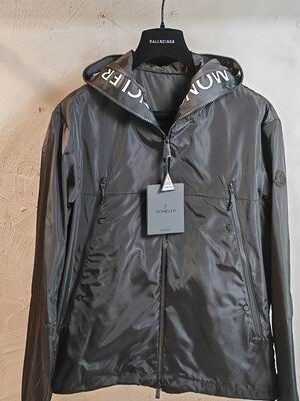 Dark grey technical hooded jacket with white branding visible inside the hood and long vertical zippered side pockets.