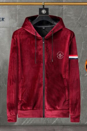 A front view of a burgundy velvet zip-up hoodie with a black zipper, thick drawstrings, a fleece-lined hood, and a white stripe detail on the sleeve, hanging in a modern room setting.