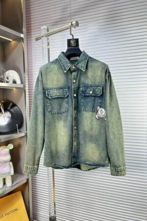 A long-sleeved denim overshirt with a vintage yellow and blue acid wash, featuring two chest pockets and a white circular logo patch on the side.